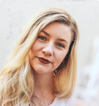 Sieviete ar gariem blondiem matiem un tumšu lūpu krāsu skatās kamerā uz gaiša fona.
