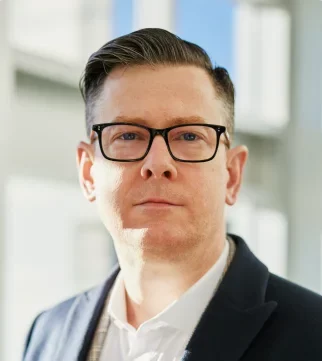 Vīrietis ar īsiem brūniem matiem un brillēm, tērpies uzvalka žaketē, stāv spilgti apgaismotā telpā.