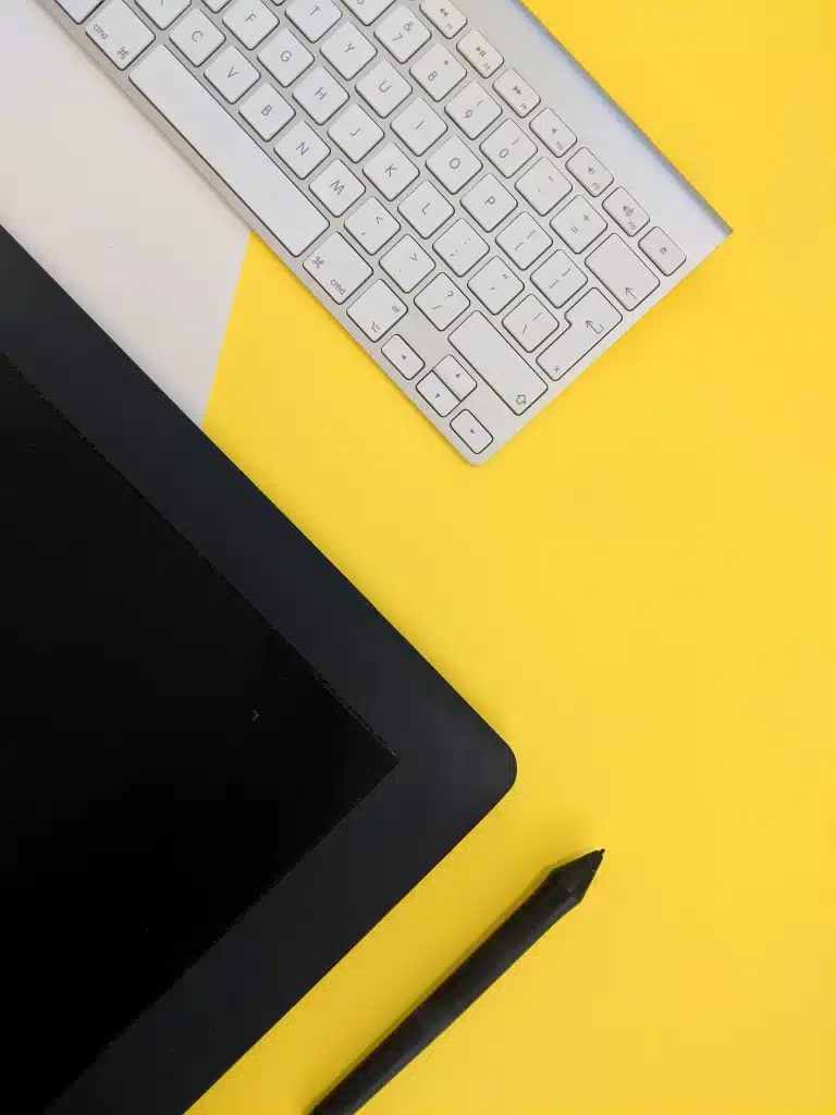 Balta tastatūra, digitālais planšetdators un pildspalva uz dzeltena un balta fona.