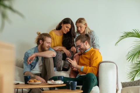 Two women and two men sitting comfortably on a sofa in a room, looking at a smartphone, a light wall in the background, a coffee table in the foreground