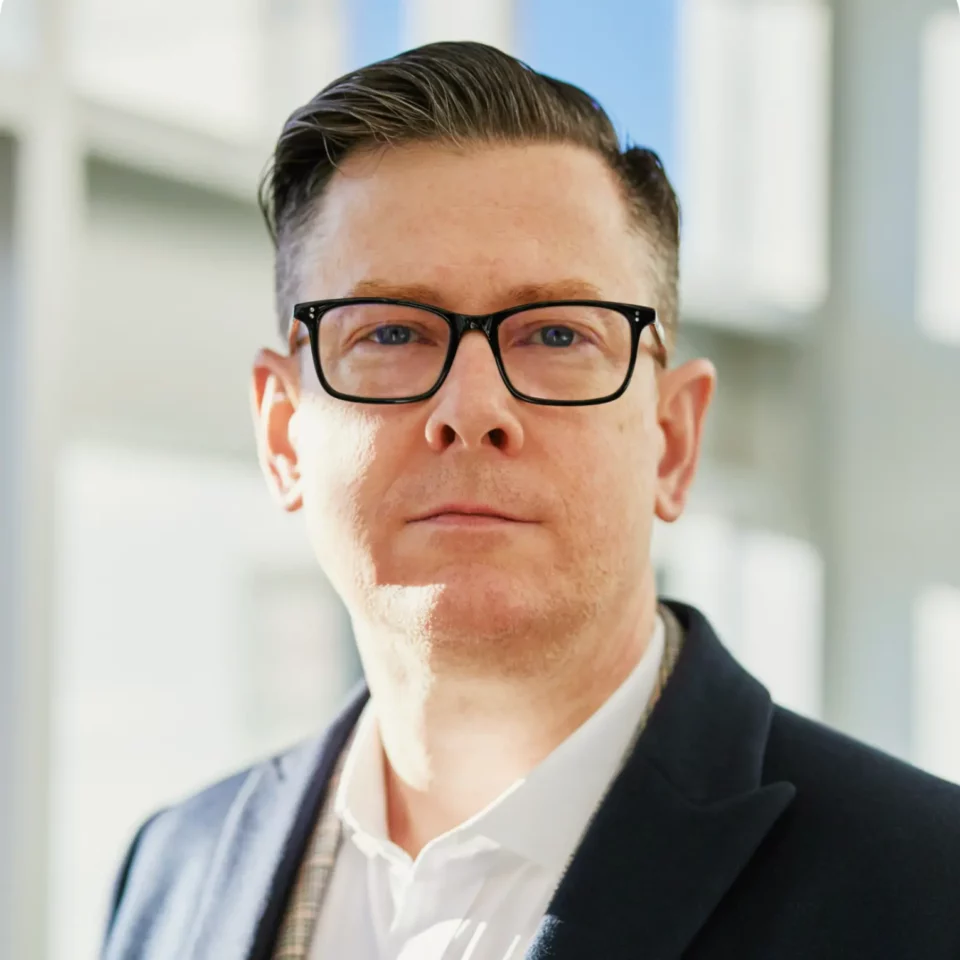 Vīrietis ar īsiem brūniem matiem un brillēm, tērpies uzvalka žaketē, stāv spilgti apgaismotā telpā.