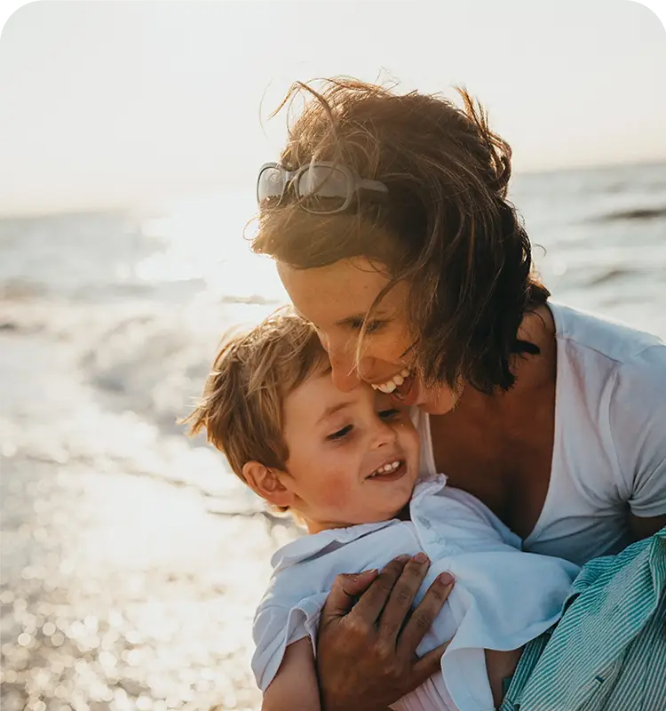 A woman hugs a smiling young child on a sunny beach, both looking happy and relaxed.