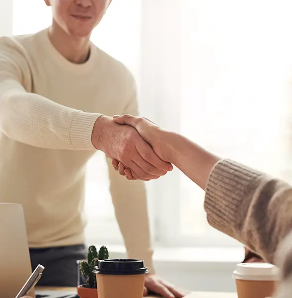 Two people shaking hands across a desk with coffee cups and a small plant in the foreground.