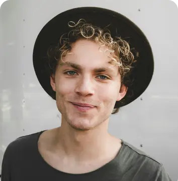 Young man with curly hair wearing a dark hat and grey shirt, smiling in front of a light background.