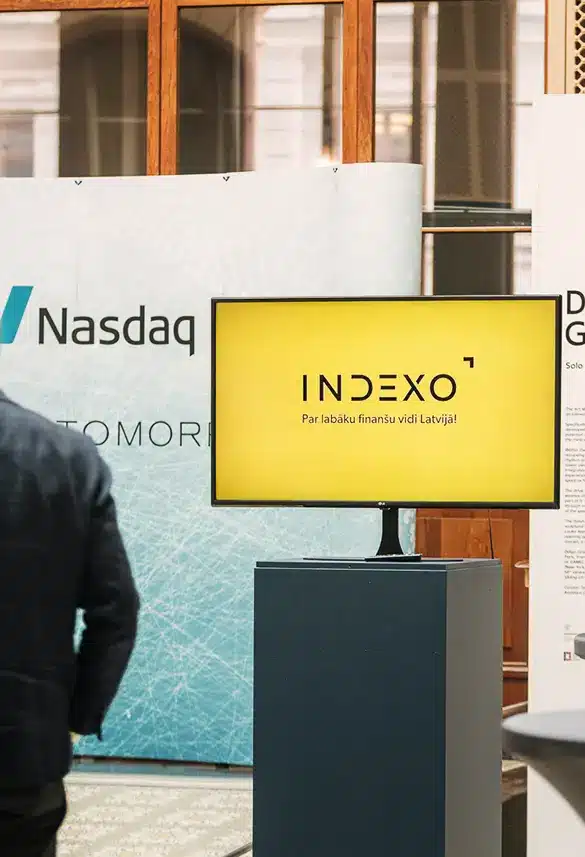 A monitor displays INDEXO next to a Nasdaq banner at an indoor event or conference.