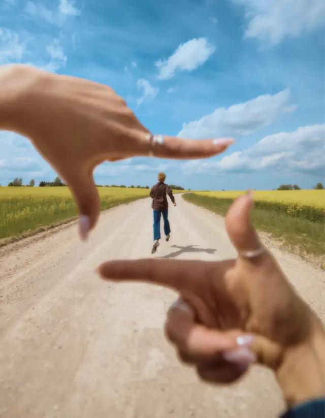 A man runs along a country road, framed between two arms in the foreground, a flowering field and blue sky in the background
